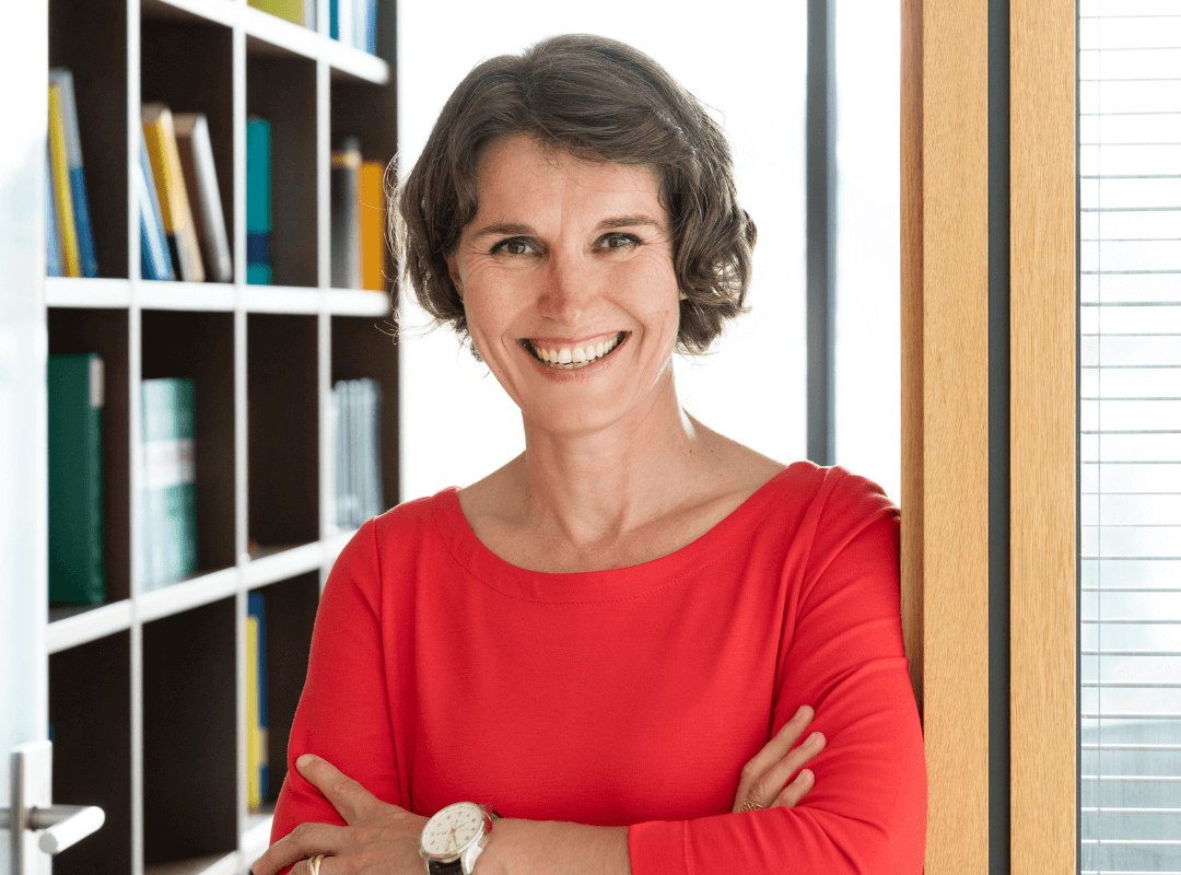 Frau mit dunklen halblanger gewellter Frisur steht im Türrahmen eines Büro-Meetingraumes. Sie trägt ein rotes Businesskleid mit halblangen Ärneln. Die Arme sind sympatisch locker vor dem körper verschränkt. Sie lächelt freundlich. Am Armgelenk trägt sie eine Uhr mit größerem Ziffernblatt und braunem Lederarmband. Am Rinfinger trägt sie Trinity. Das Etuikleid hat einen oval-runden Ausschnitt sowie Gürtel und Seitentaschen und ist von dem deutschen Business-Kleid-Label Vitoria Johansson, dass alle Kleider in Deutschland designed und fertigt.