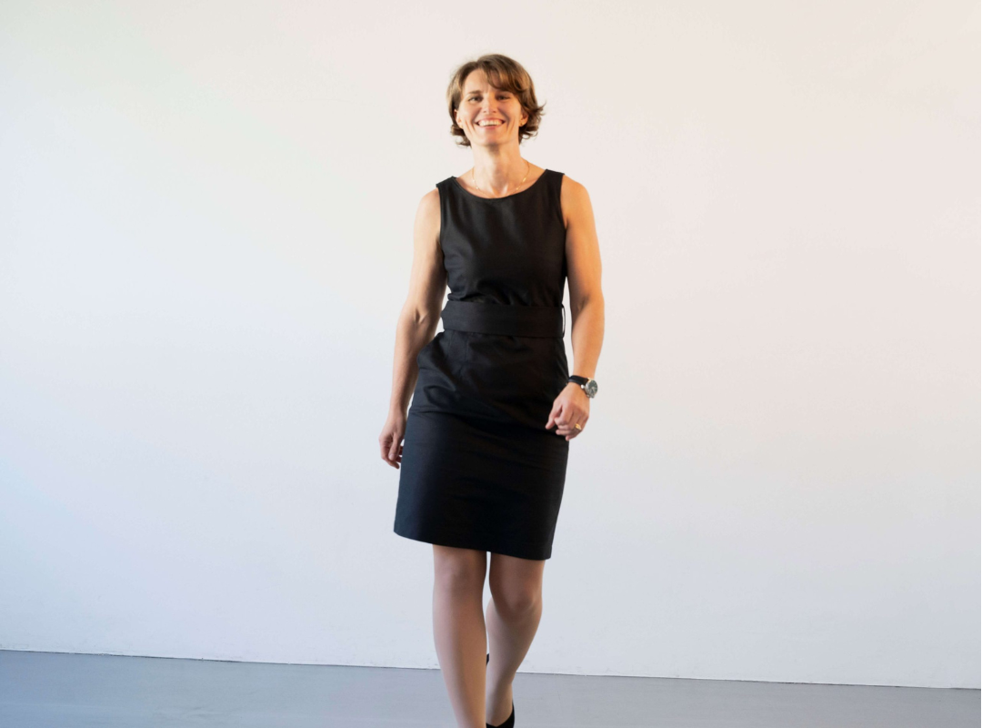 Frau vor hellblauem Hintergrund in einem schwarzen figurbetonten Etuikleid mit Träger und ovalen Halsausschnitt. Das Businesskleid hat einen breiten Gürtel, ebenfalls in schwarzer Farbe. Außerdem hat das Kleid an beiden Seiten Eingrifftaschen, die man aber nicht sieht. Sie läuft selbstbewusst und dynamisch. Zum Kleid trägt sie klassische schwarze Highheels.