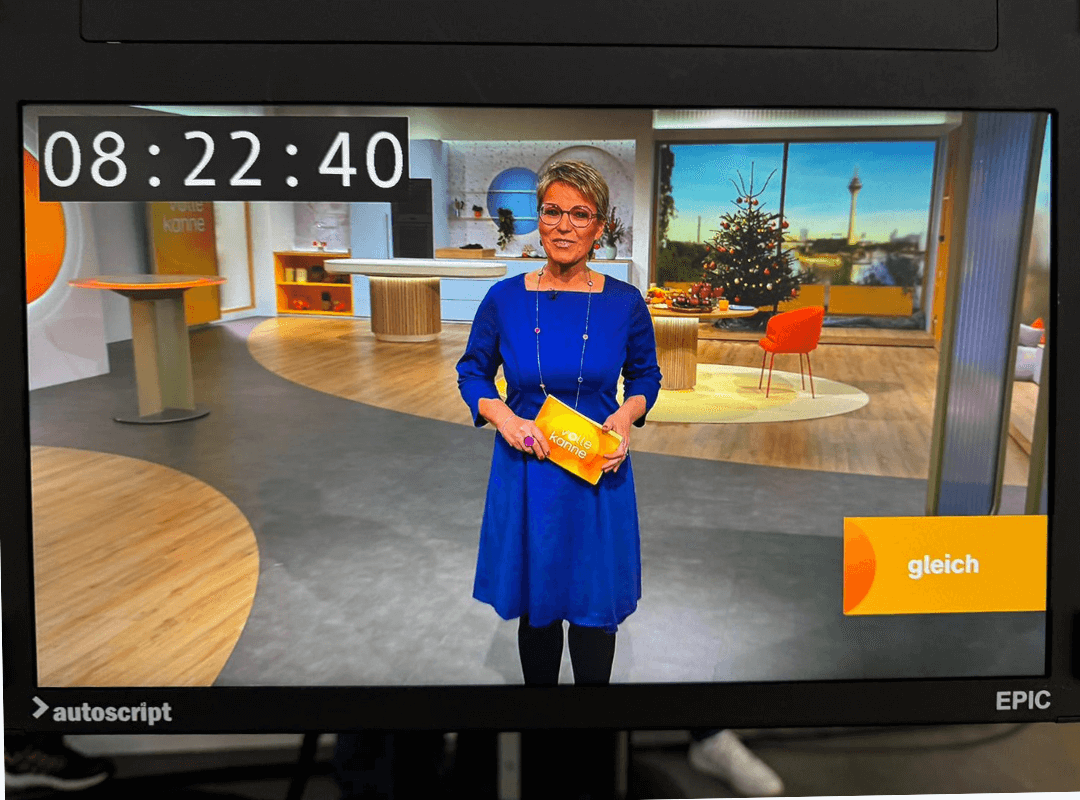 Fernsehmoderatorin im Fernsehstudio der ZDF-Sendung Volle Kanne. Sie trägt eine schicke Kurzhaarfrisur und eine Brille. Das Fernsehstudio in warmen Farbtönen ist im Hintergrund und im Vordergrund die Moderatorin in einem jeansblauen Kleid mit eckigen Ausschnitt und knielangem Rock, sowie halblangen Ärmeln. Die Kleidfarbe ist sehr gut abgestimmt zu den Studiofarben. Das Kleid ist das Modell Judith vom deutschen Businesskleid-Label Vitoria Johansson, welches seine Kleider in Deutschland designed und fertigt.