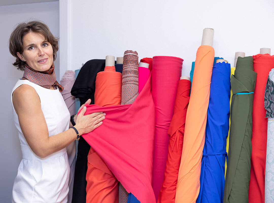 Frau in einem eleganten weißen Etuikleid und passendem Tuch steht vor einer Auswahl von Stoffen in unterschiedlichen Farben. Den Stoff in der Farbe himbeere zeigt sie hervor.