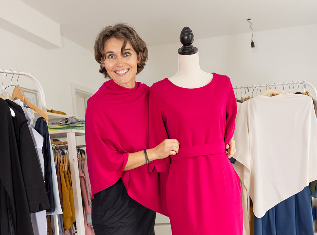 Die Frau trägt ein schwarzes knielanges Kleid und darüber einen Poncho in fuchsia. Die Modepuppe neben ihr trägt ein pinkes Etuikleid mit passendem Gürtel.